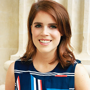 Young woman with shoulder-length auburn hair, smiling, wearing a blue patterned sleeveless top, standing outdoors with stone columns in the background.