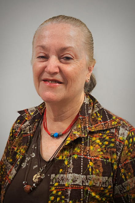 An older woman with gray hair pulled back, wearing a colorful patterned jacket and layered necklaces, standing against a plain gray background.