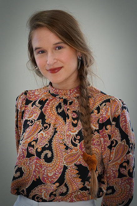A young woman with long wavy hair in a side braid, wearing a paisley patterned blouse with puffy shoulders, smiling at the camera against a plain background.