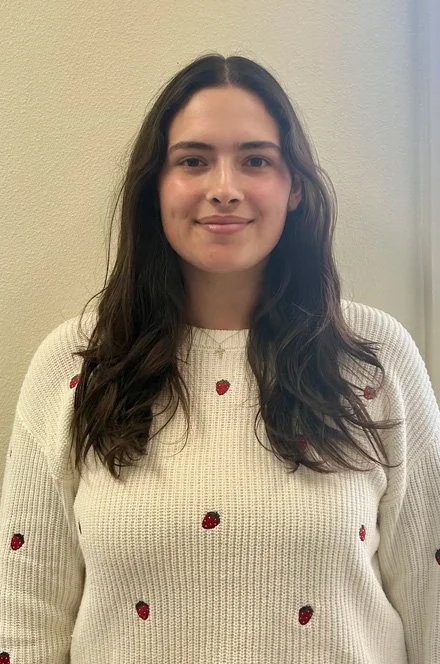 A young woman with long dark hair standing indoors in front of a neutral-colored wall, wearing a cream sweater with small strawberry prints.