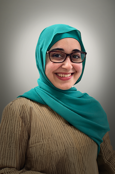 A woman wearing a teal hijab, glasses, and a beige ribbed sweater smiling at the camera