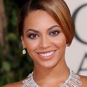 Close-up of a smiling woman with light brown hair, wearing diamond earrings and a pearl necklace, against a blurred background.