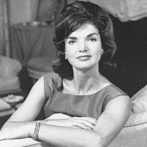 A woman with dark, shoulder-length hair, wearing a sleeveless dress, sitting with her arms crossed.