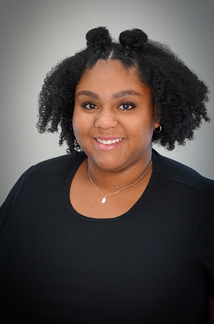 Portrait of a young woman with curly hair styled in puffs, wearing a black shirt and jewelry, smiling against a plain gray background.