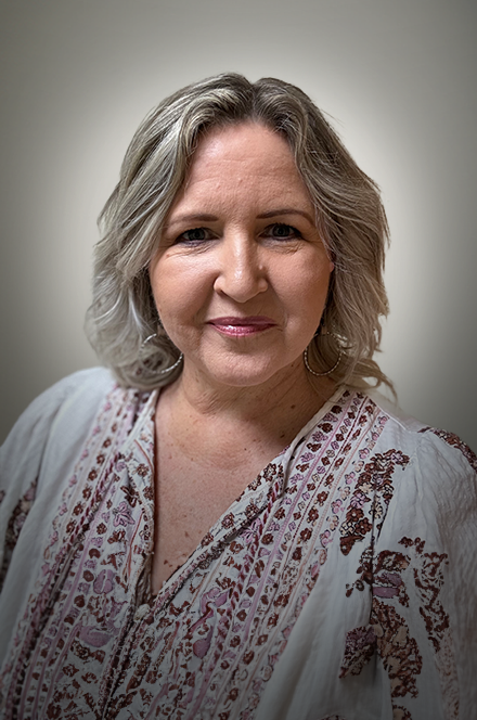 A middle-aged woman with shoulder-length gray hair and light skin, smiling slightly, wearing a white blouse with embroidered patterns and large hoop earrings, standing in front of a plain neutral-colored wall.