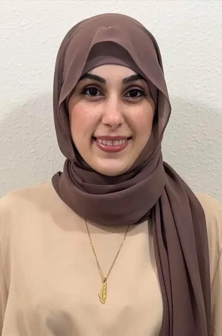 A woman wearing a light brown hijab and beige top, smiling at the camera, with a gold necklace that has a leaf pendant, standing against a plain light-colored wall.