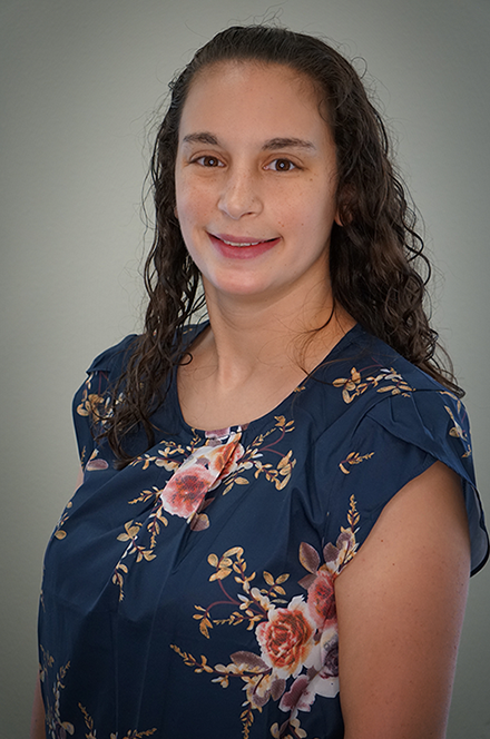 A woman with long, curly brown hair smiling, wearing a navy blue floral blouse, standing against a plain gray background.
