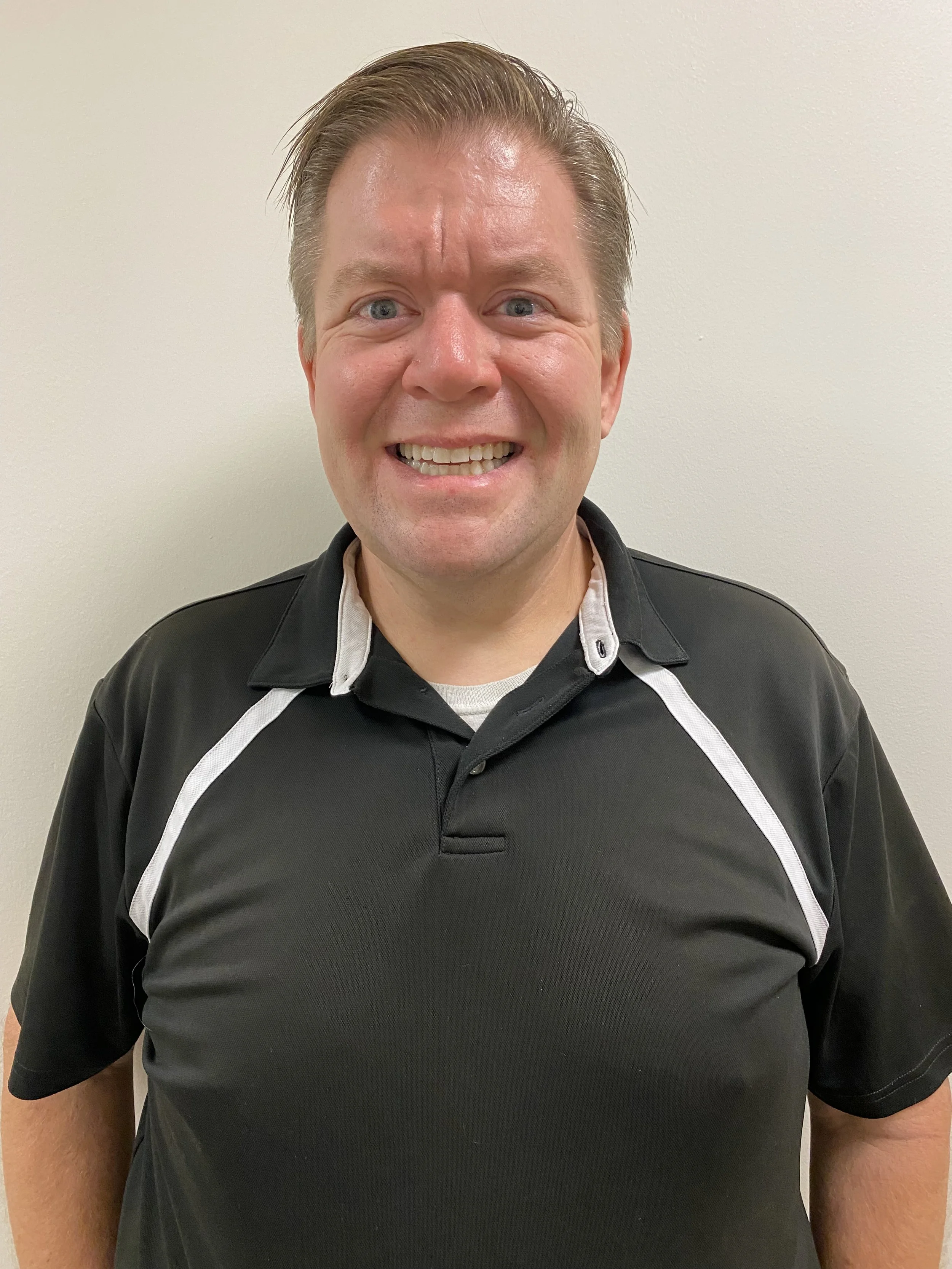 A man smiling and looking at the camera, wearing a black polo shirt with white accents, standing against a plain white wall.