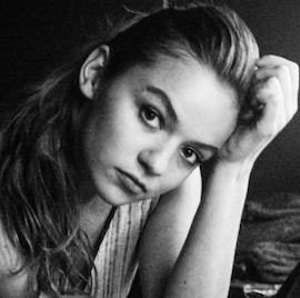Black and white portrait of a young woman with long hair, resting her head on her hand and gazing at the camera.