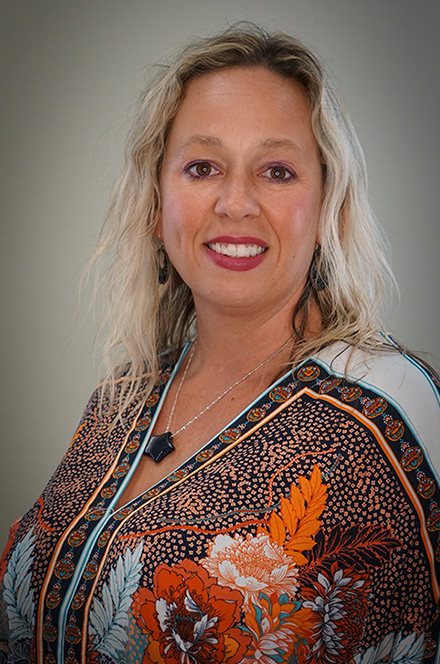 A smiling woman with blonde curly hair wearing a patterned blouse with floral designs and a necklace, standing against a plain gray background.