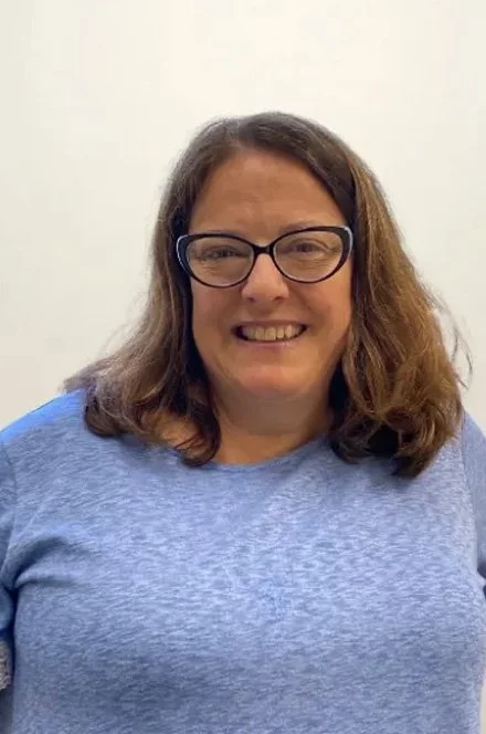 A woman with brown hair, black glasses, and a blue shirt smiling at the camera against a plain white background.