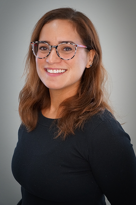 A woman with shoulder-length brown hair, wearing glasses with clear and pink frames, smiling at the camera, dressed in a black long-sleeve top, standing against a plain light gray background.