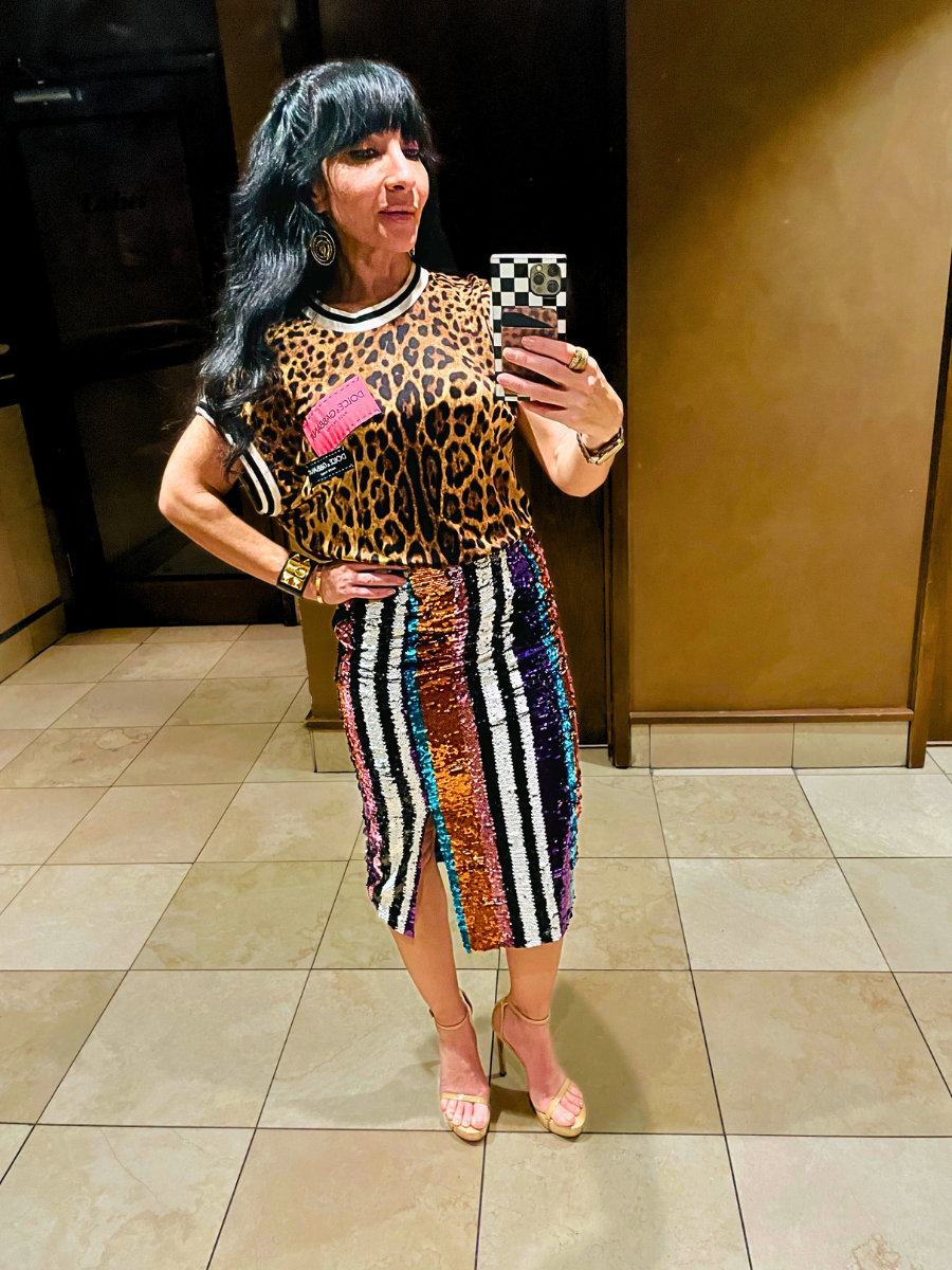 Woman taking a mirror selfie in a fashionable outfit, wearing a leopard print top with a tag and a colorful sequined skirt with a slit, beige high heels, and accessories including earrings, bracelet, and rings.