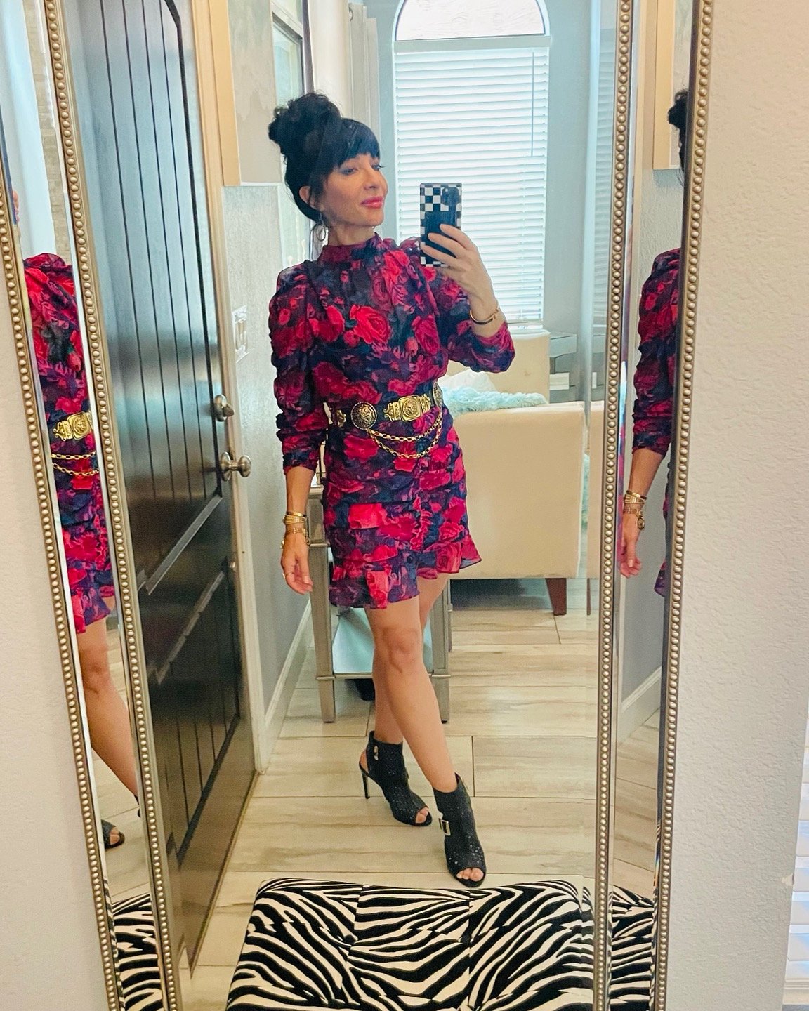 A woman taking a mirror selfie in a fashionable outfit with a floral red and black dress, black high heels, and gold accessories, in a room with a large window with blinds and a zebra print rug.