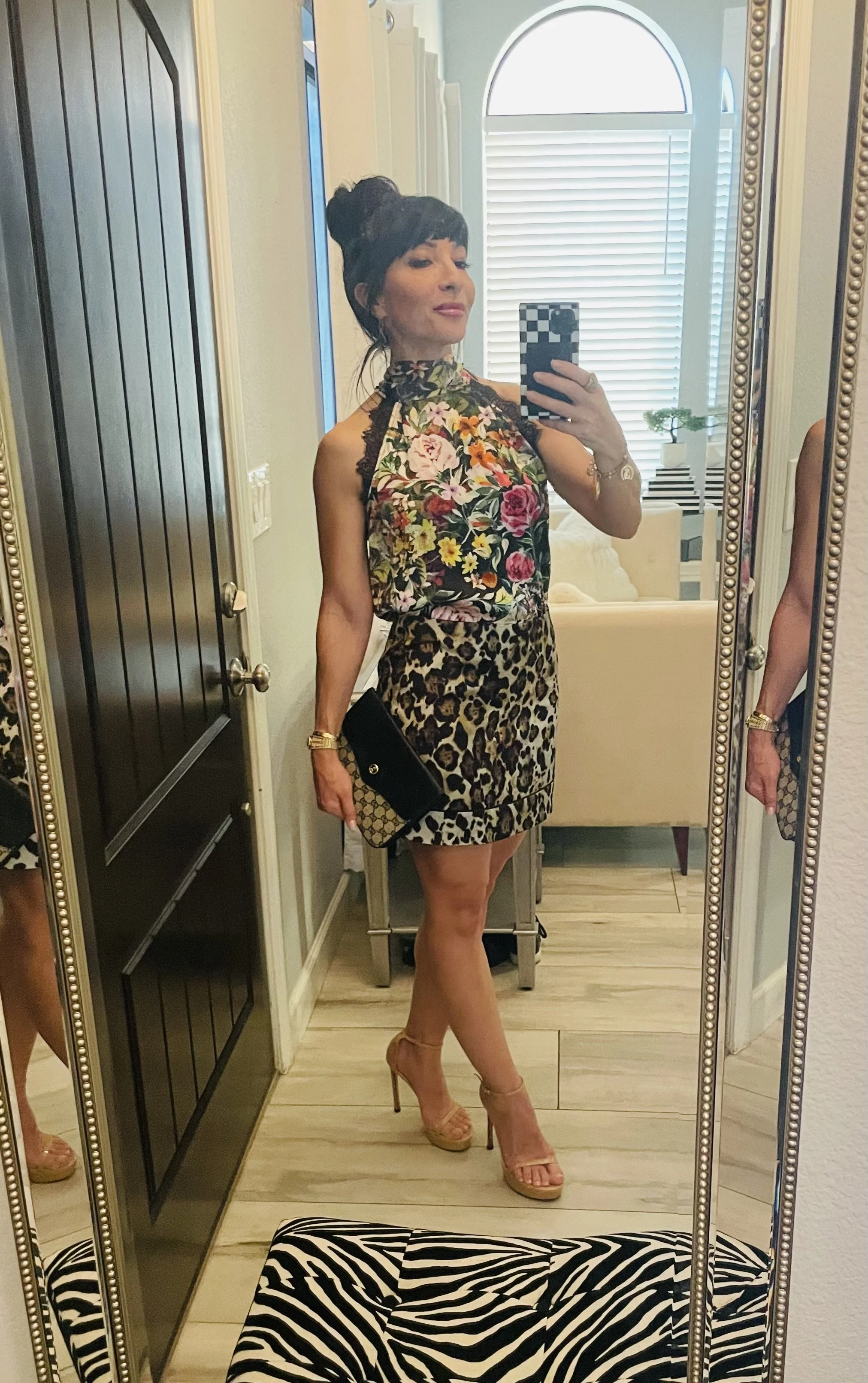A woman taking a mirror selfie dressed in a floral top with a high neckline and a leopard print skirt. She is wearing beige high-heeled sandals and holding a black clutch. The background shows a bright room with blinds covering large windows, a beige sofa, and a small table with a plant on it.