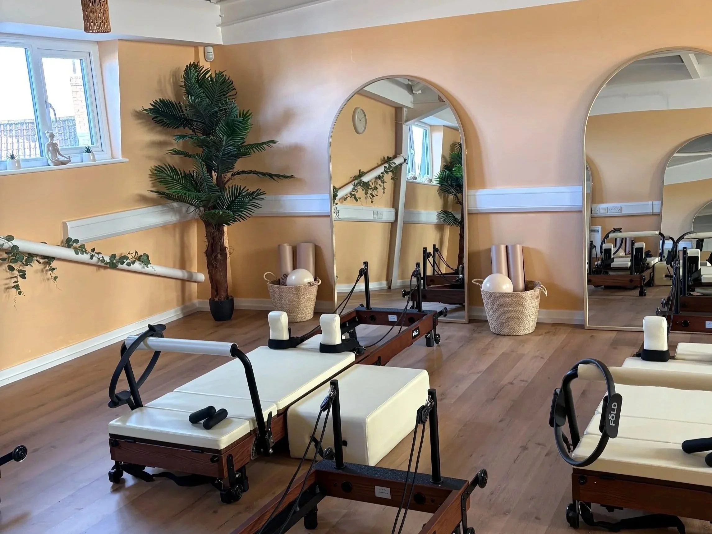 A room with yellow walls and wooden floors, decorated with potted plants, mirrors, exercise equipment, and sunlight coming through the windows.