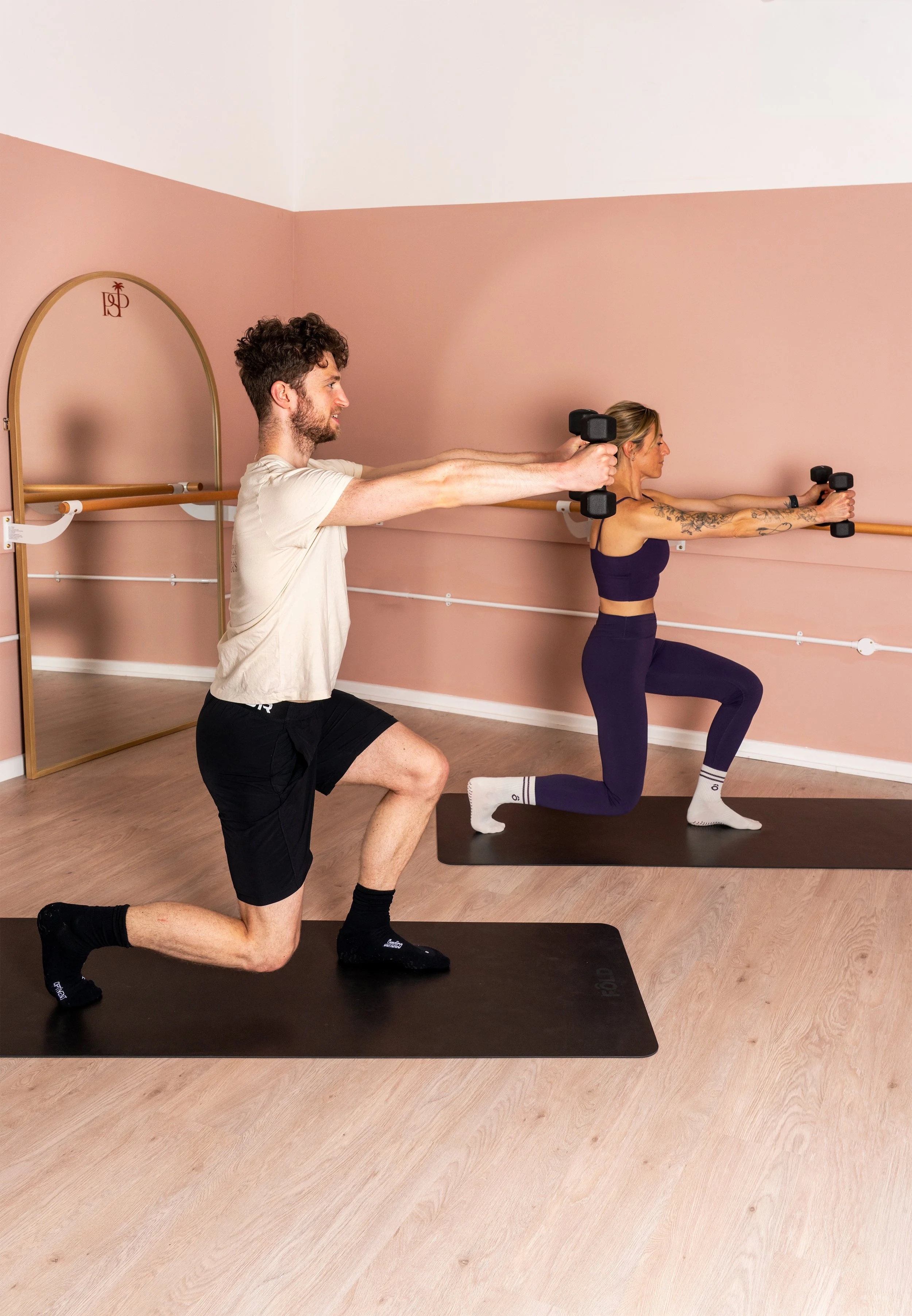 A man and woman participating in a fitness class in a studio. The woman is doing a lunge with dumbbells, while the man is kneeling behind her, assisting. They are on black exercise mats, and the studio has light wooden floors and pink walls with a ba