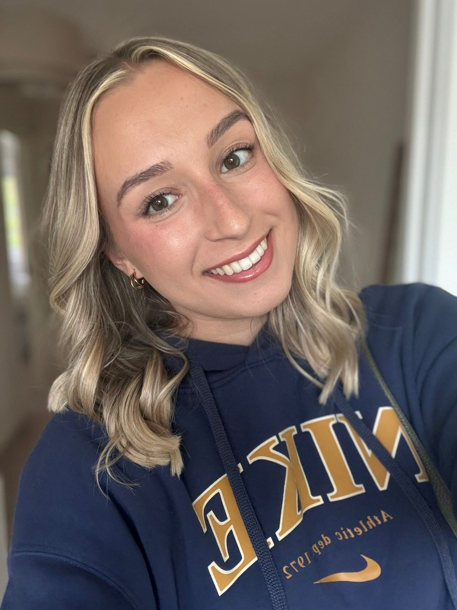 A young woman with blonde wavy hair smiling in a navy blue hoodie with gold lettering.