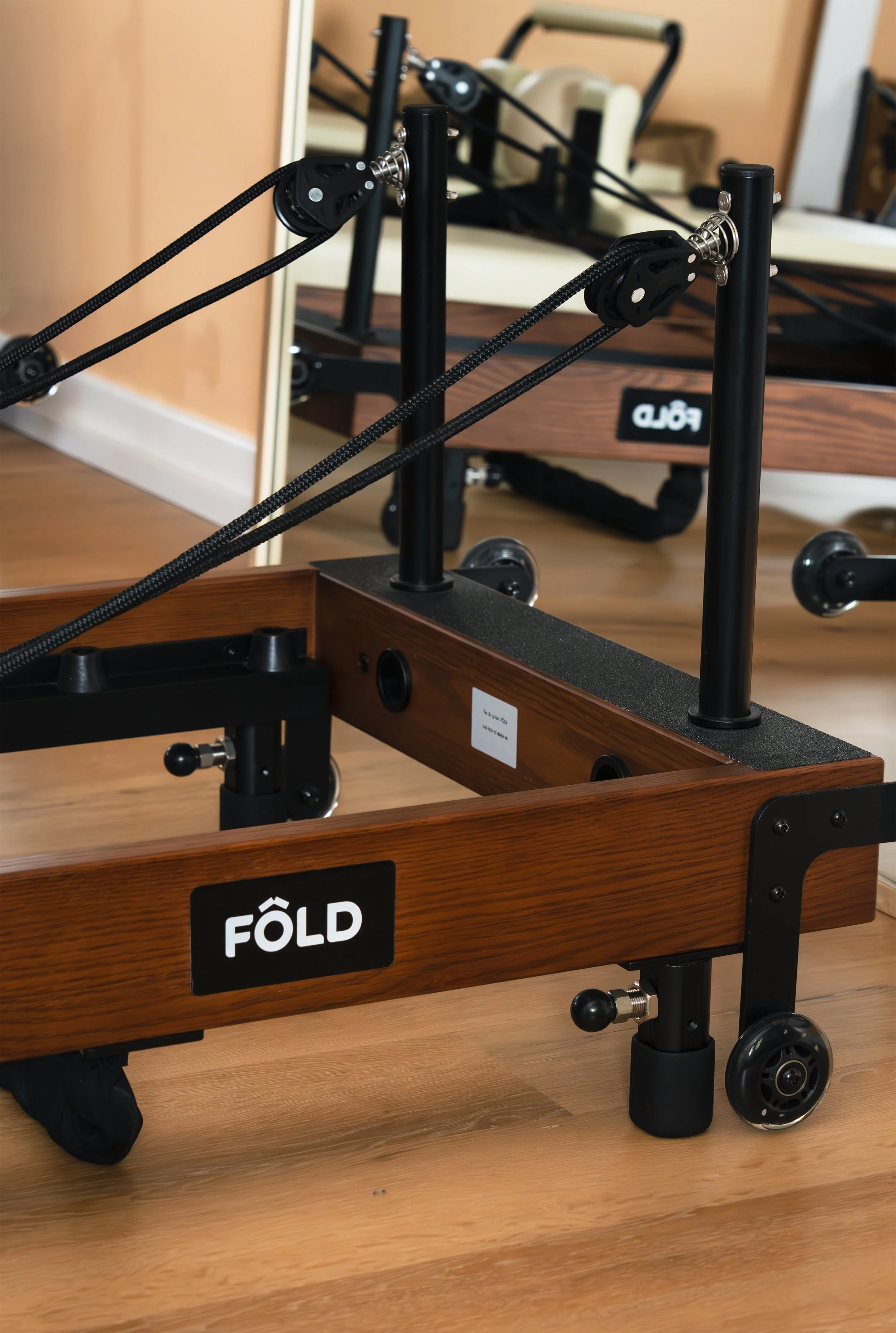A wooden folding cart with black metal frame, labeled 'FÖLD,' situated on a wooden floor next to a mirror, with a beige couch in the background.