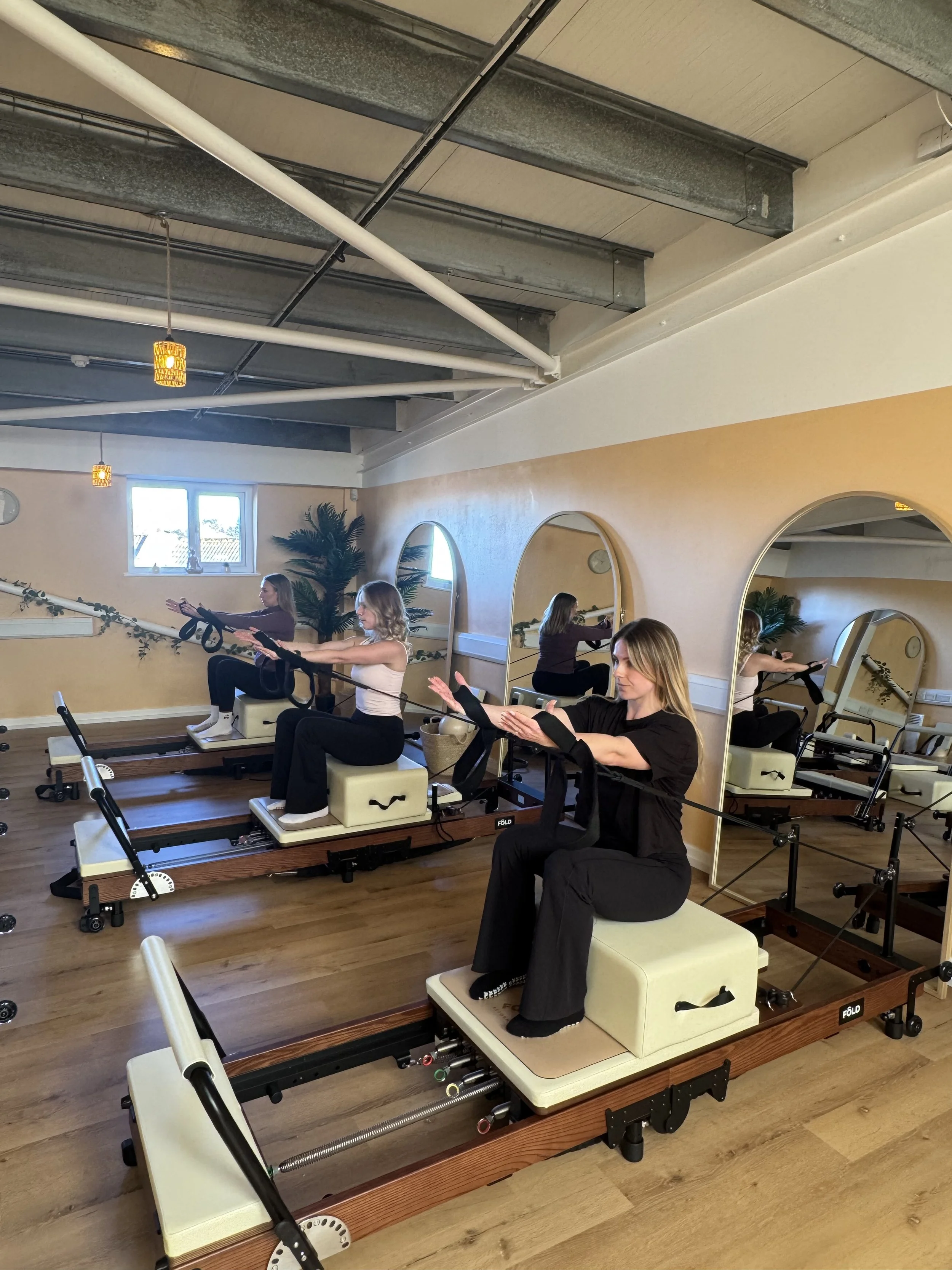 Four women practicing Pilates on reformer machines in a fitness studio with mirrors, plants, and pendant lighting.