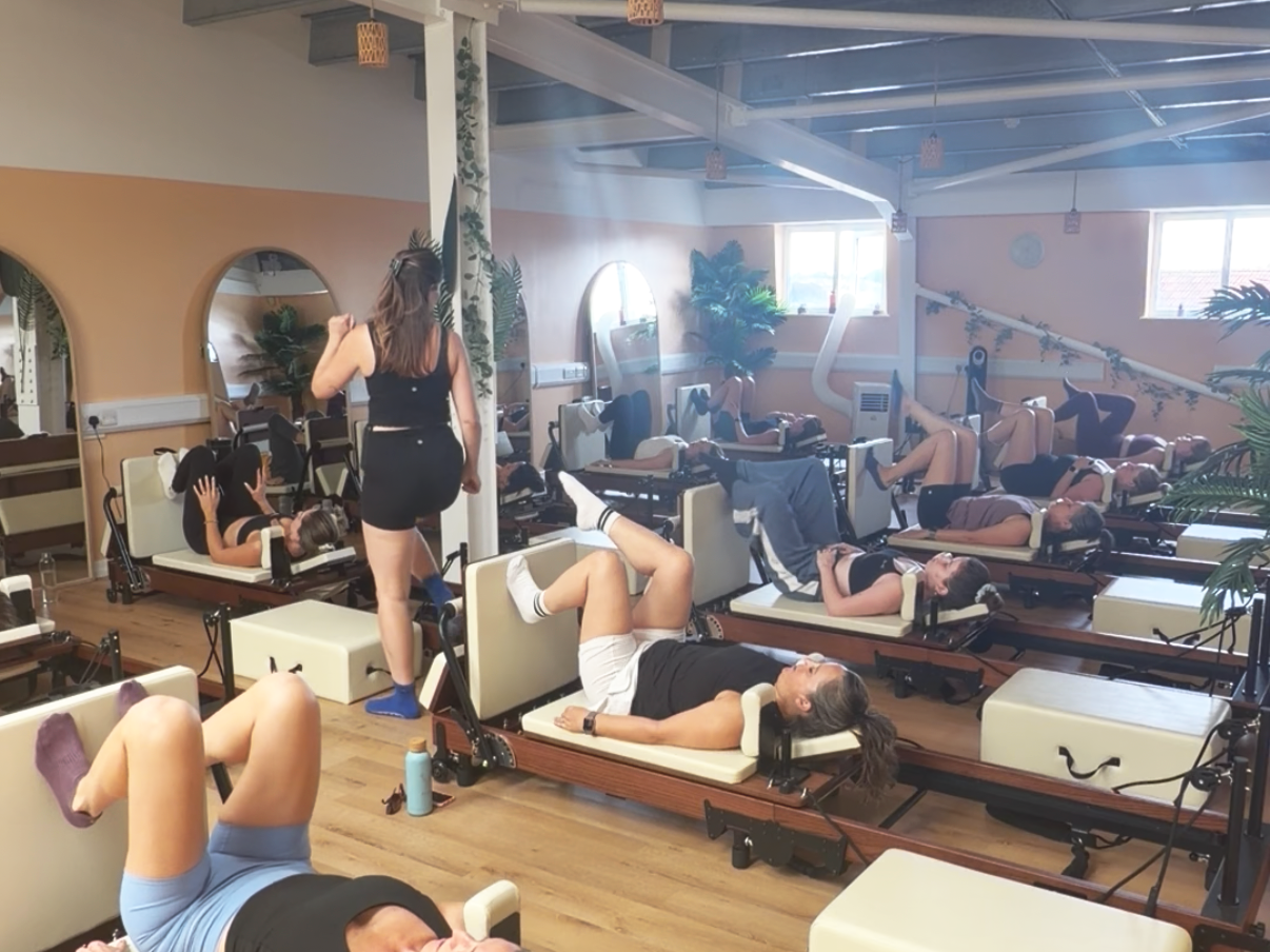 A Pilates studio with several people lying on reformer machines, some doing exercises, and an instructor walking among them, with mirrors and plants in the background.