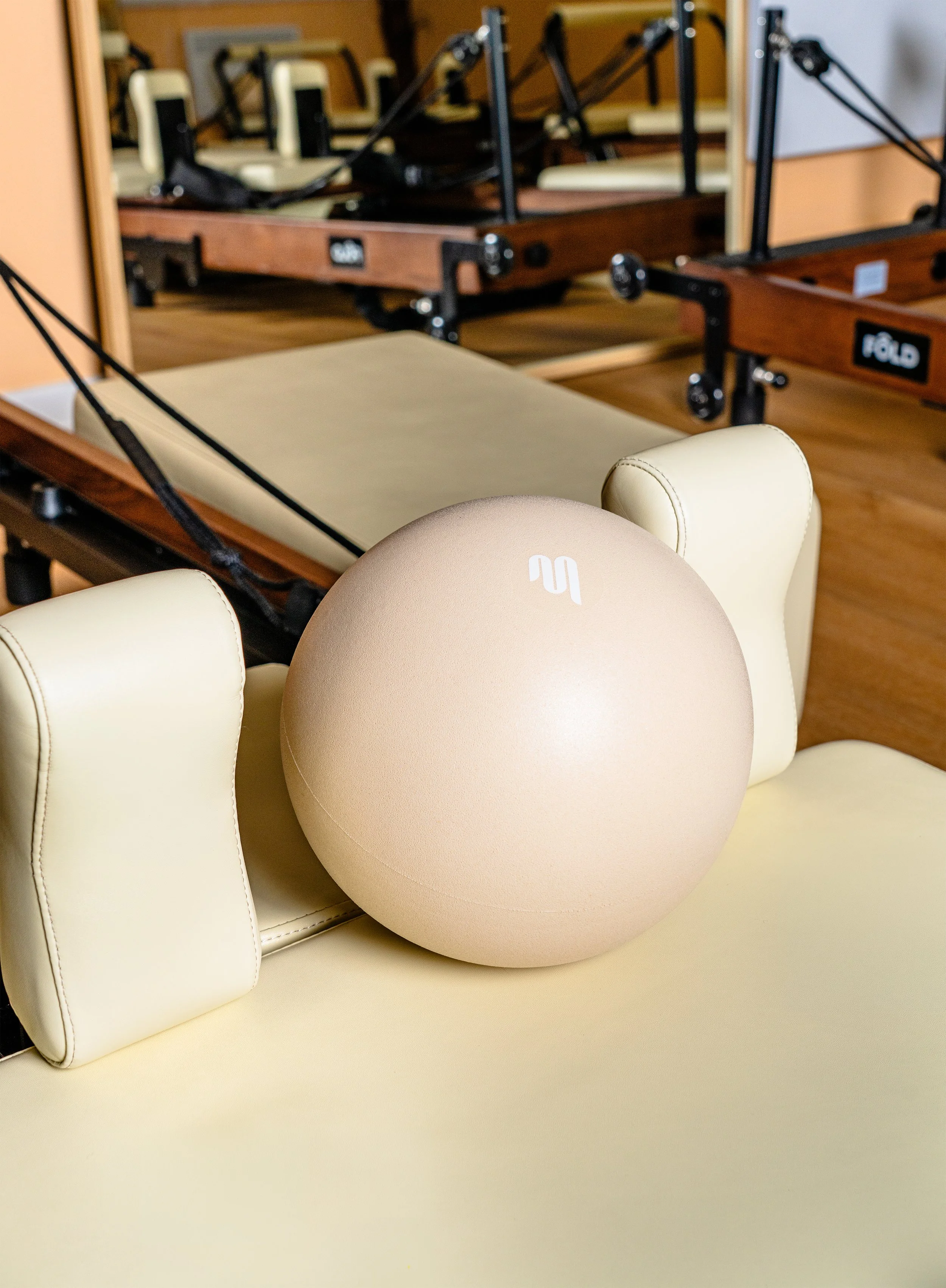 A beige exercise ball with a white logo resting on a cream-colored couch, with a mirror, a wooden table, and chairs in the background.