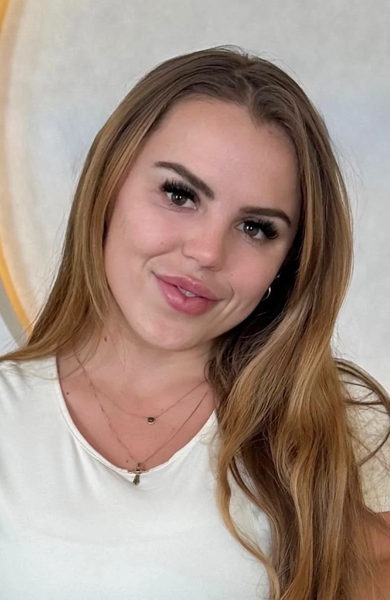 A young woman with long, wavy auburn hair, wearing a white top and layered necklaces, smiling slightly, standing in front of a plain light-colored wall.