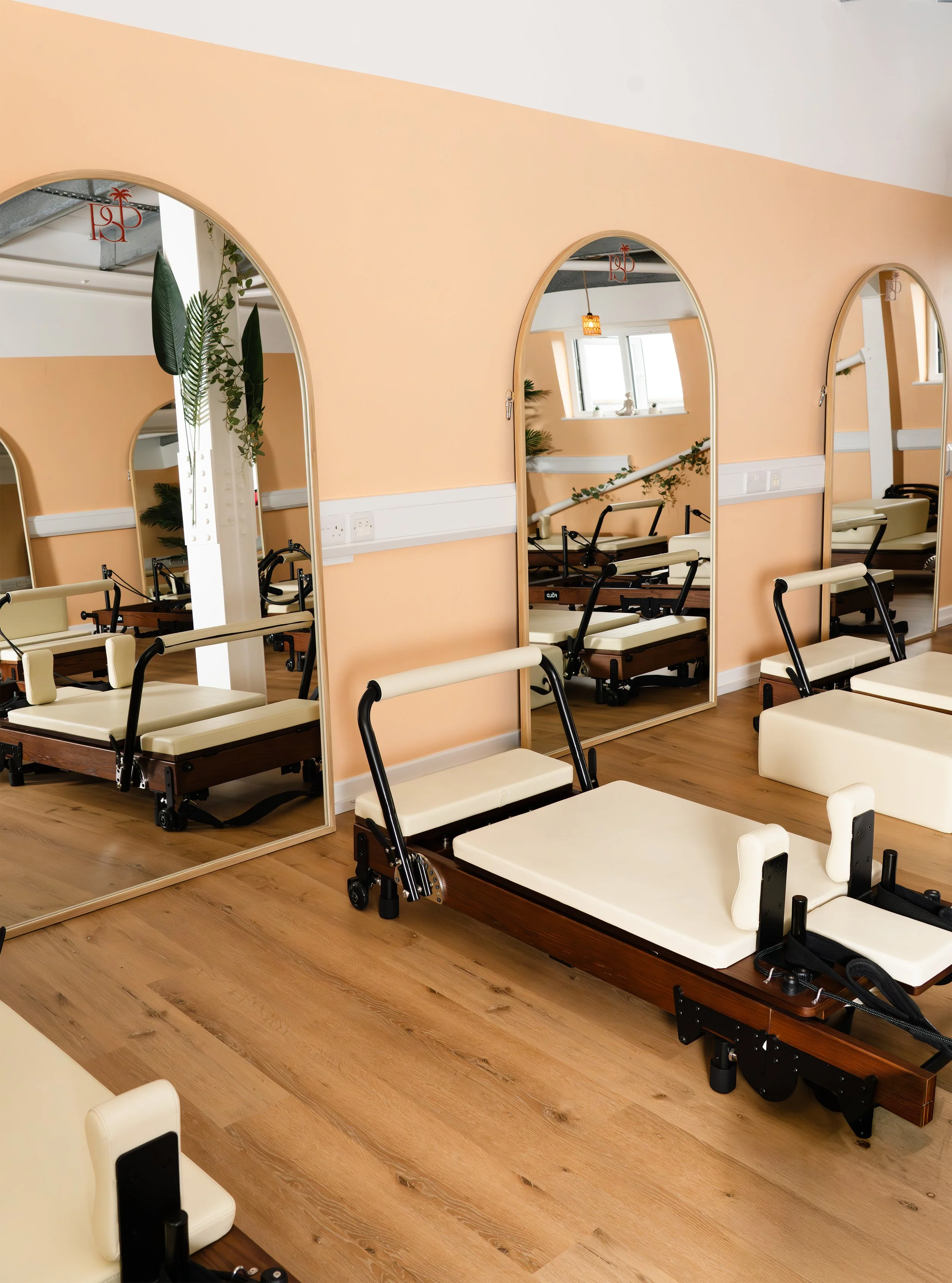 Room with Reformer Pilates beds and mirrors, decorated with plants and warm-colored walls.