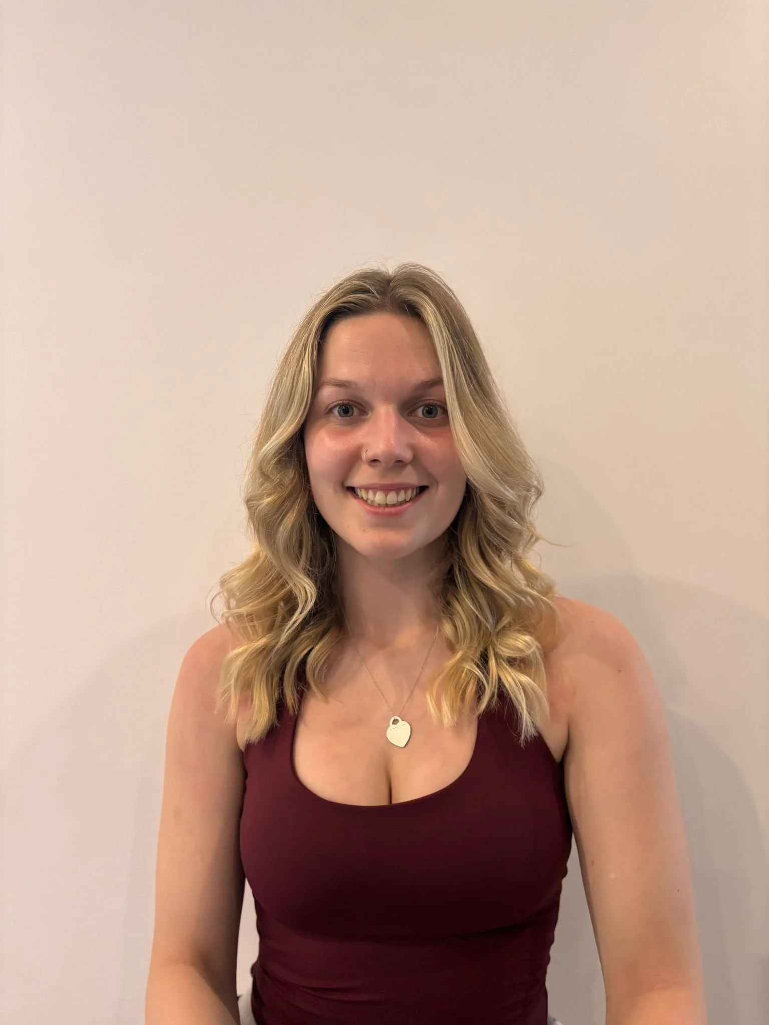 A young woman with shoulder-length blonde wavy hair, smiling, wearing a maroon sleeveless top and a necklace with a heart-shaped pendant, standing against a plain white wall.