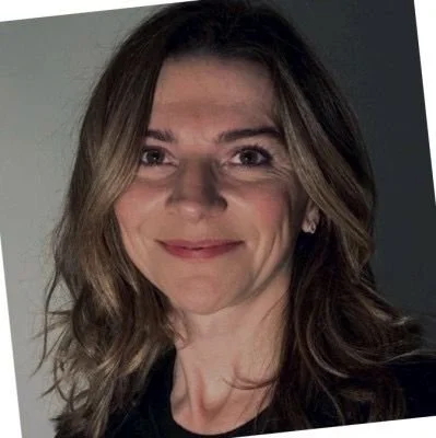 A woman with shoulder-length brown hair and a slight smile, wearing a black top, standing indoors against a neutral background.