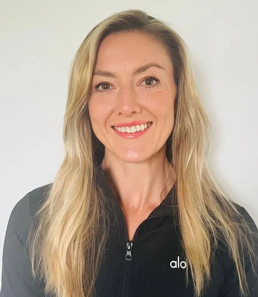 Portrait of a woman with long blonde hair smiling, wearing a black zip-up top with the word 'alo' on it, standing against a plain white background.