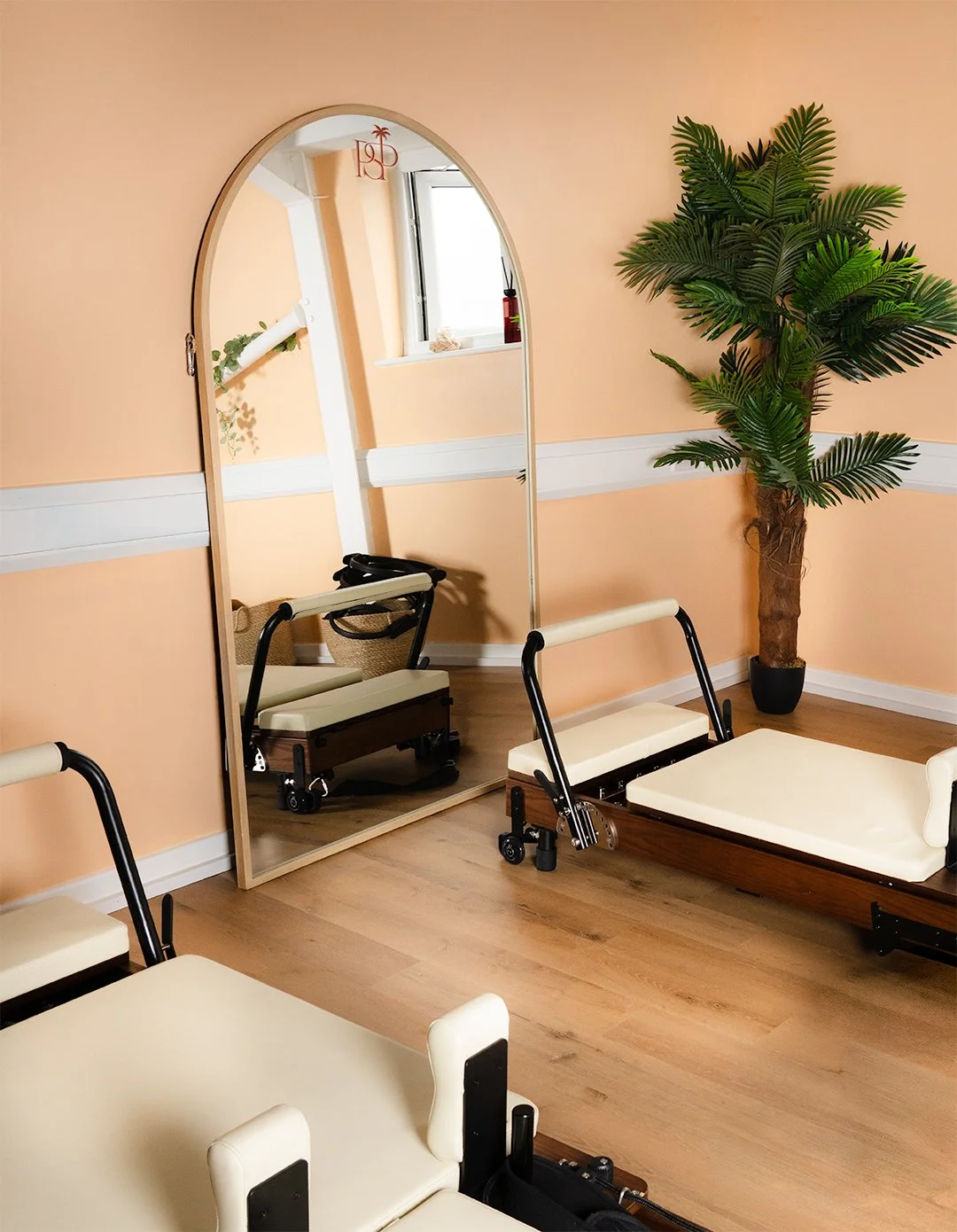 A Pilates studio room with a large mirror, a potted artificial palm tree, a window, and a Pilates reformer machine on the floor with cushioned supports, all set against peach-colored walls with white trim.