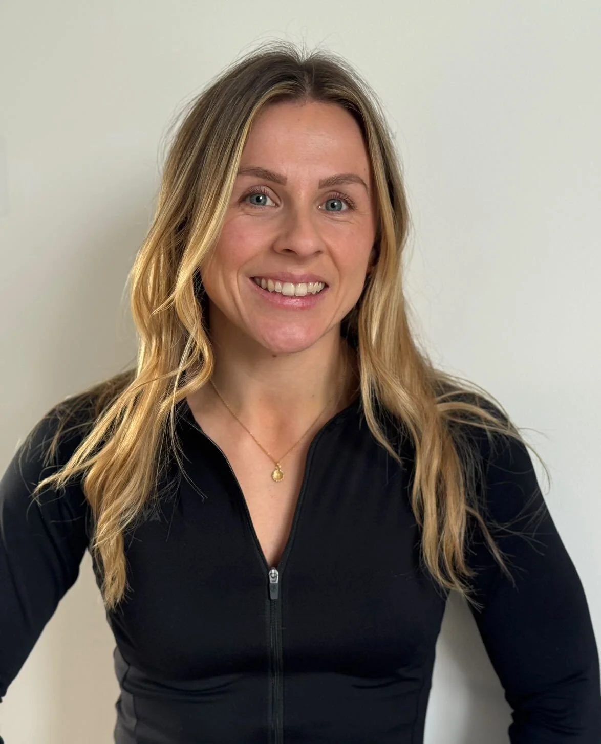 A woman with long, wavy blonde hair smiling at the camera, wearing a black zip-up athletic jacket and a gold necklace, against a plain white wall.