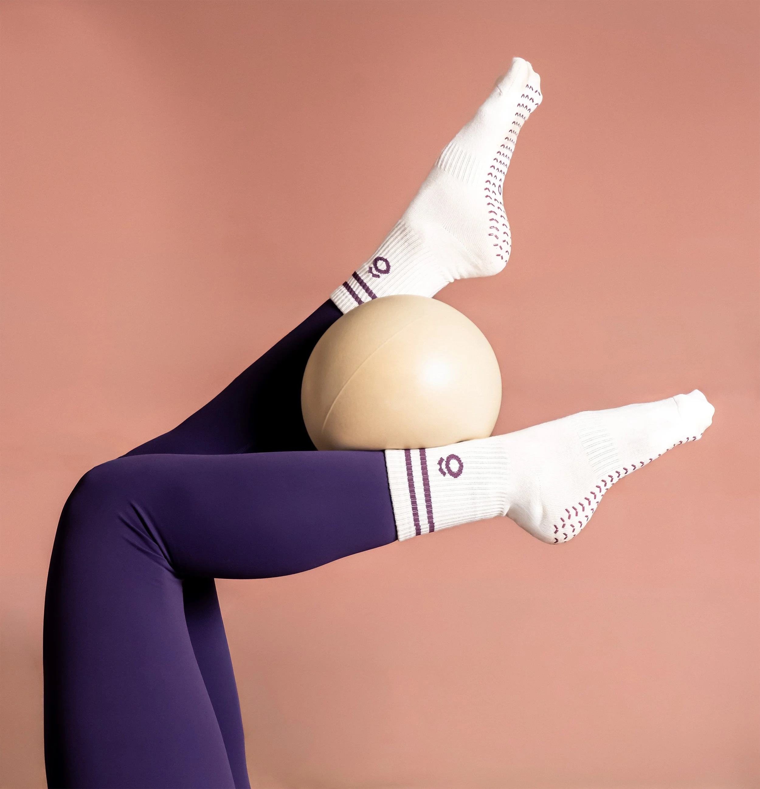 Person lying on their back with their legs raised, wearing white socks and black leggings, next to a beige exercise ball on a pink background.