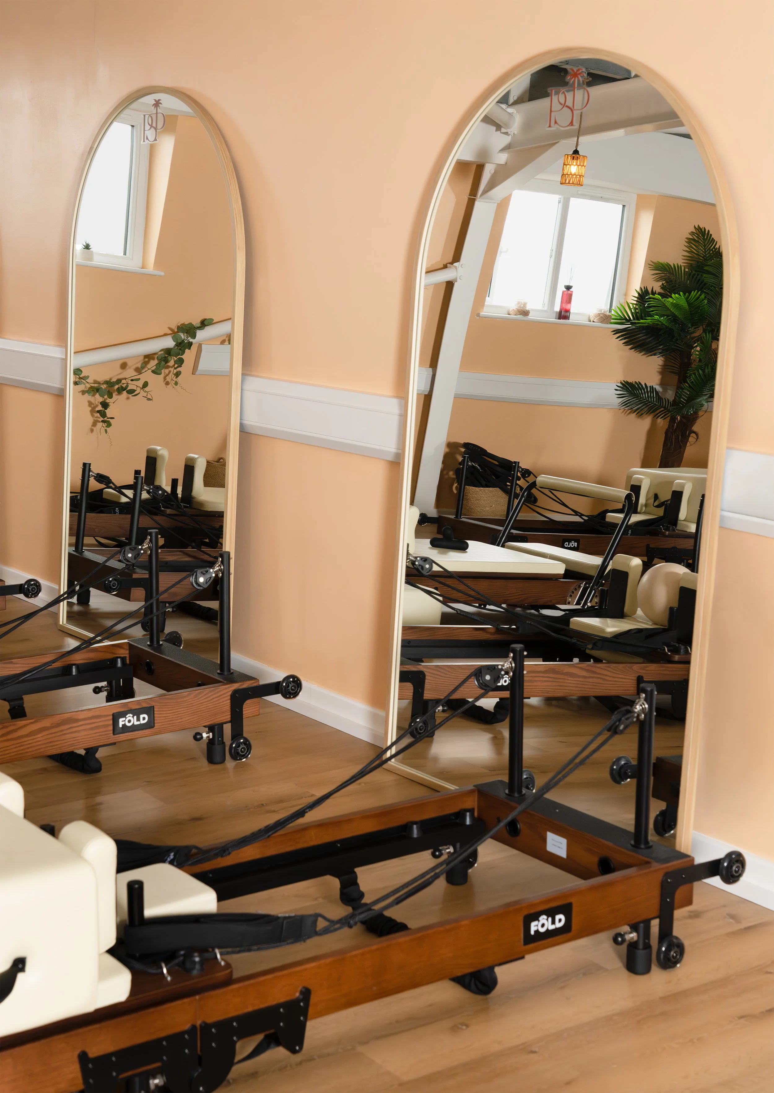 A Pilates studio with peach-colored walls and wooden flooring, featuring multiple Pilates reformer machines arranged in rows, a large mirror on the wall reflecting the room, and a window with some decorative items on the sill, including a plant, a re