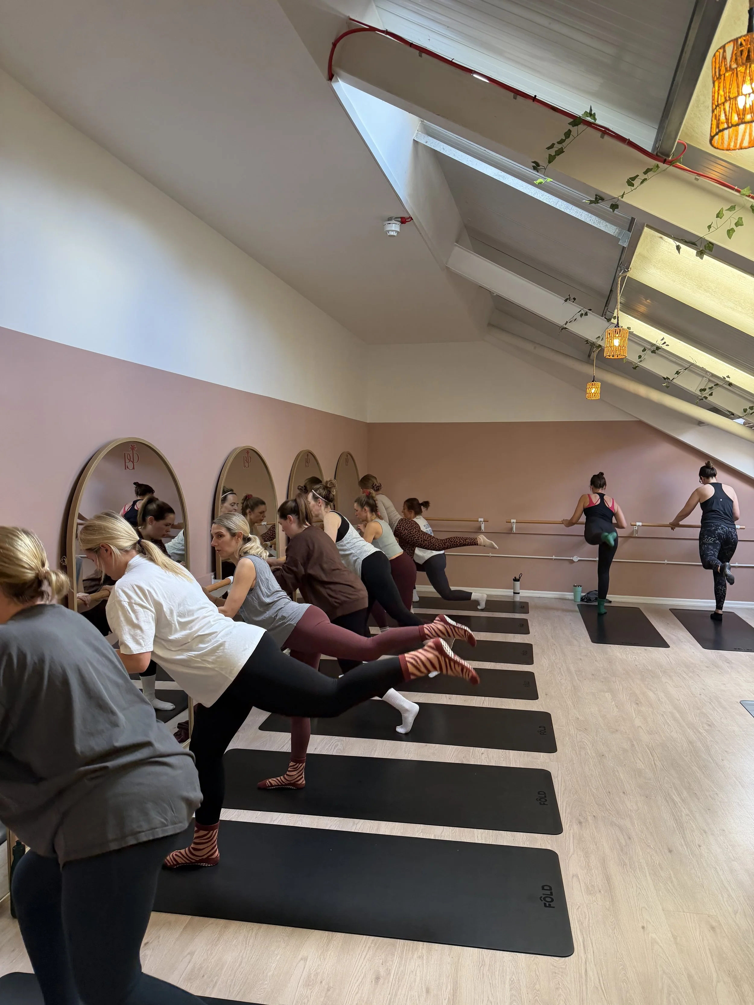 Women practicing yoga in a studio with mirrors, and an angled ceiling with skylights and hanging lights.