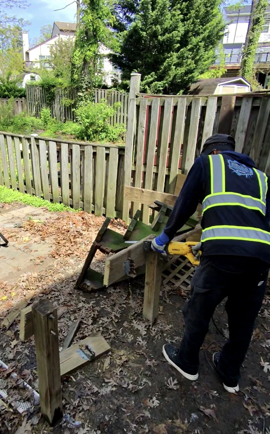 A person wearing a black jacket with yellow reflective stripes and gloves is using a power saw to cut a wooden fence post in a backyard. Leaves are scattered on the ground, and there is a partially dismantled wooden fence with a garden area and trees in the background.