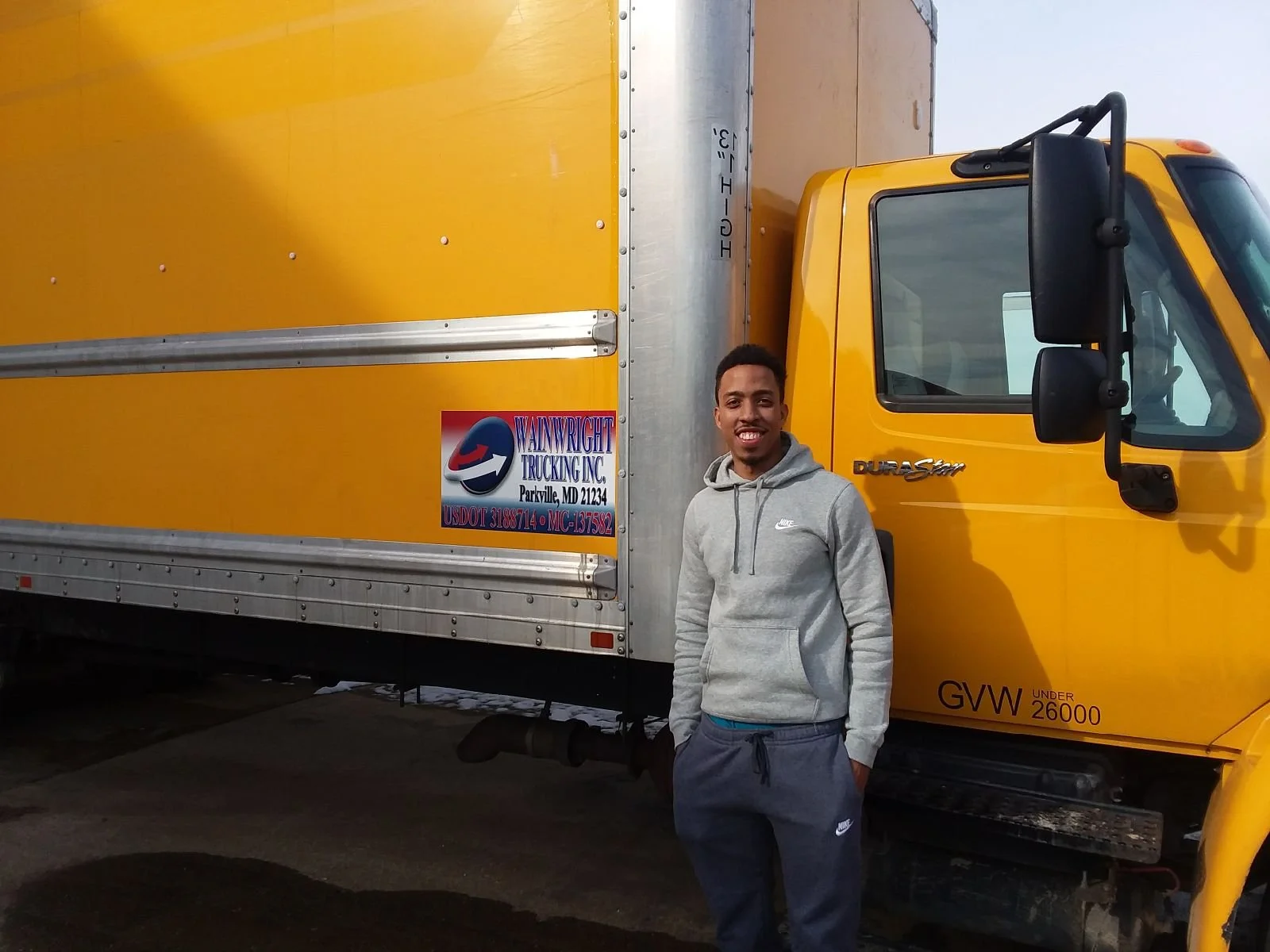 A young man standing in front of a yellow delivery truck, smiling at the camera. The truck has a logo for Walwright Trucking Inc. and is parked outdoors.