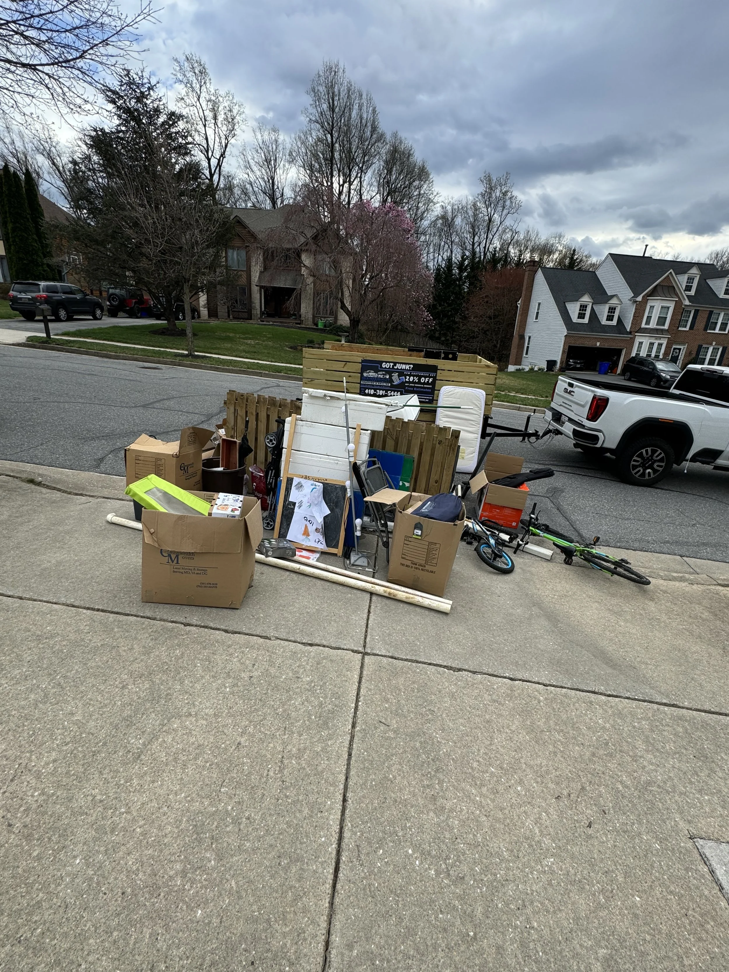 A pile of discarded household items, including boxes, a bicycle, and various furniture, placed on the sidewalk in front of a residential neighborhood on a cloudy day.