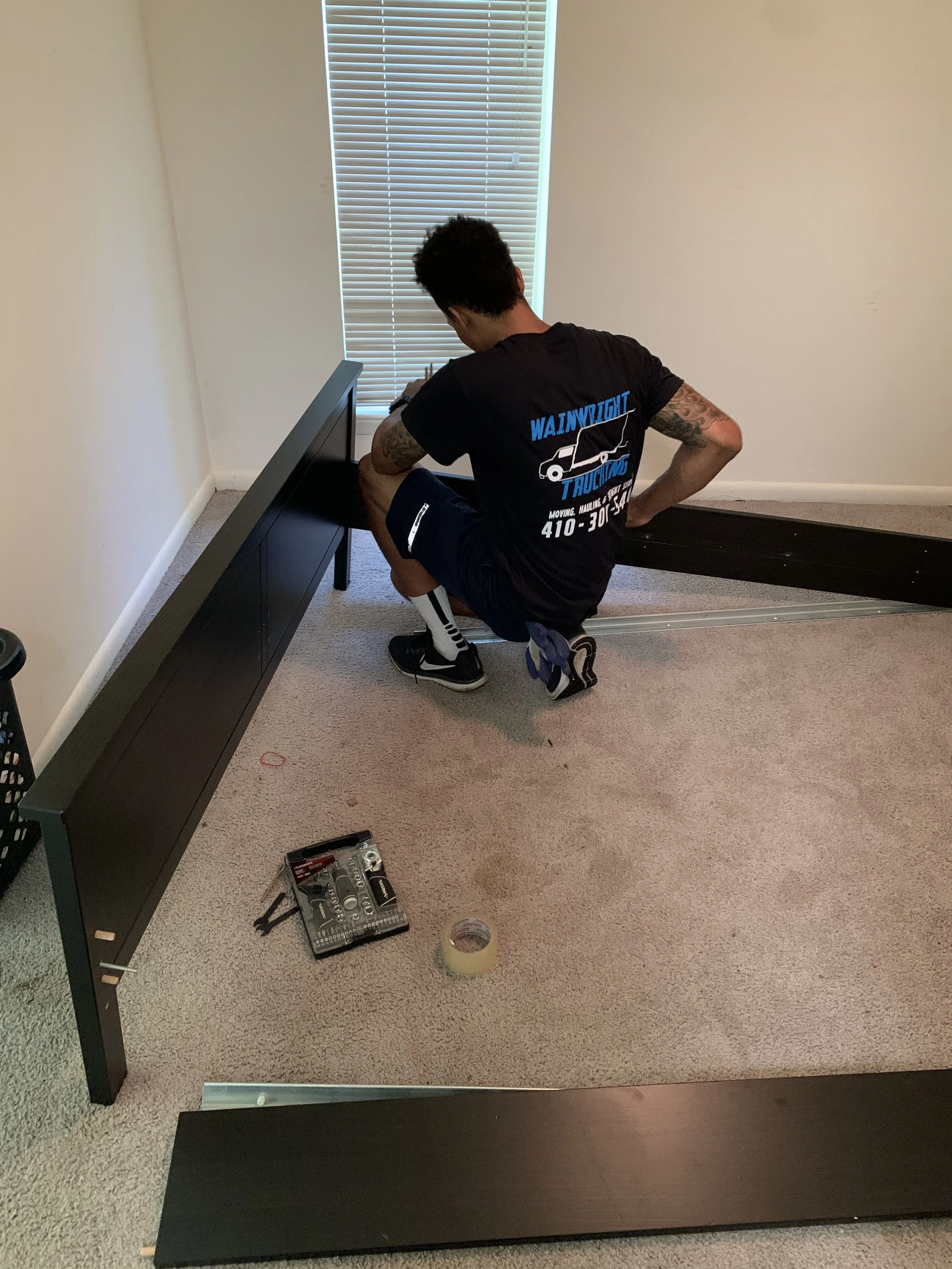 A man kneeling on a carpeted floor assembling black furniture with tools and measuring tape nearby, near a window with closed blinds.