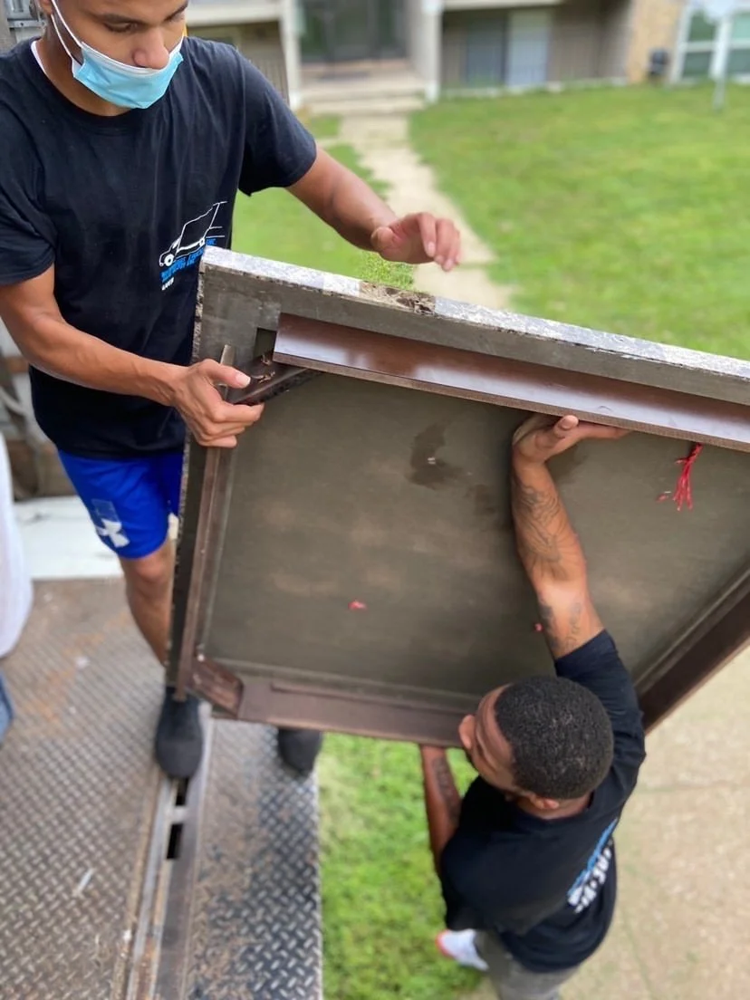Two men are working together to lift a large, flat object, possibly a piece of furniture or equipment, during daylight outdoors. Both men are wearing dark shirts and the man underneath has a tattooed arm, while the one above is wearing a face mask and blue shorts.