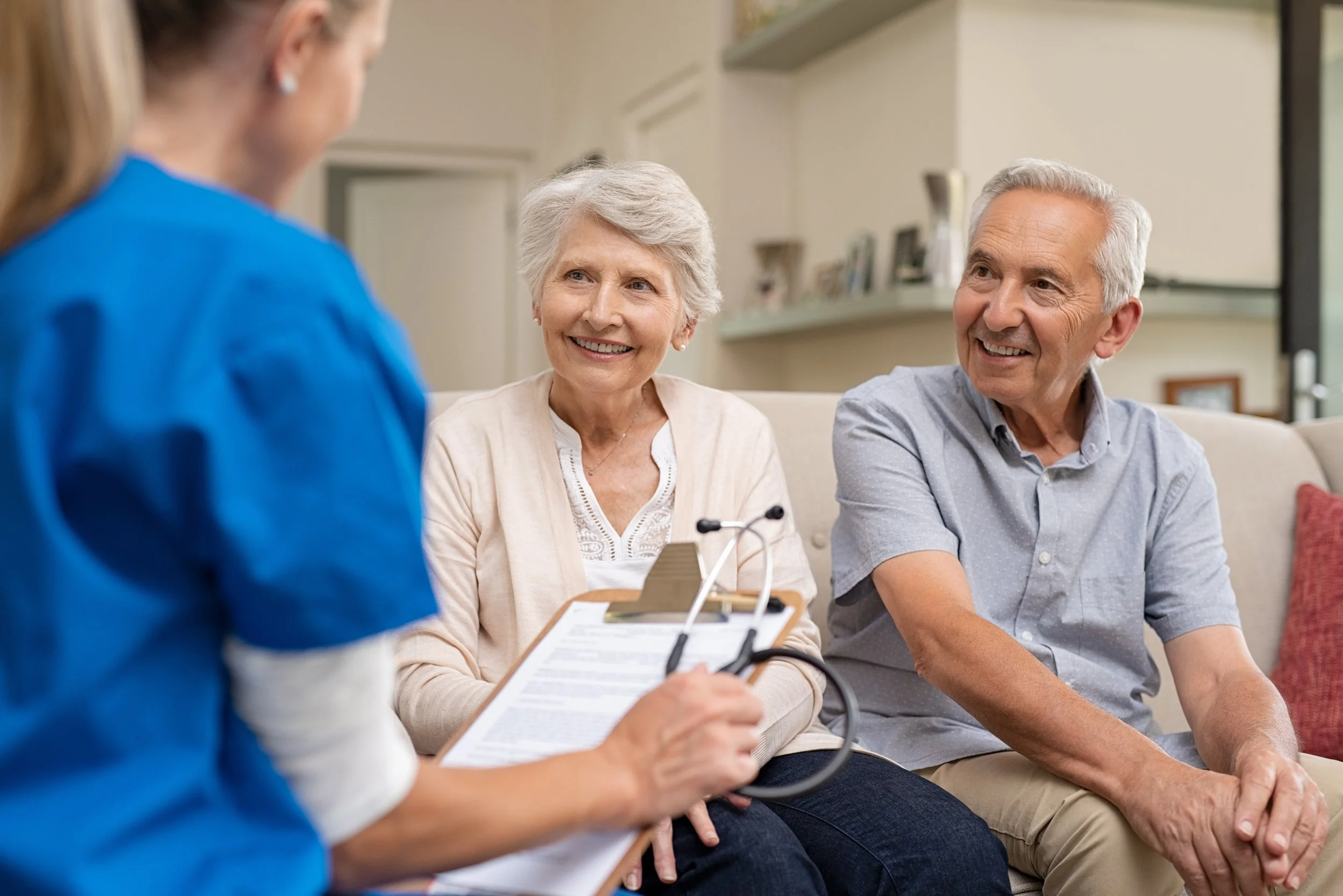 An elderly couple sitting on a beige couch in a living room, talking to a healthcare professional who is taking their blood pressure. The woman is smiling, wearing a white cardigan, and the man is smiling, wearing a light blue shirt. The healthcare professional is holding a clipboard and a blood pressure cuff.