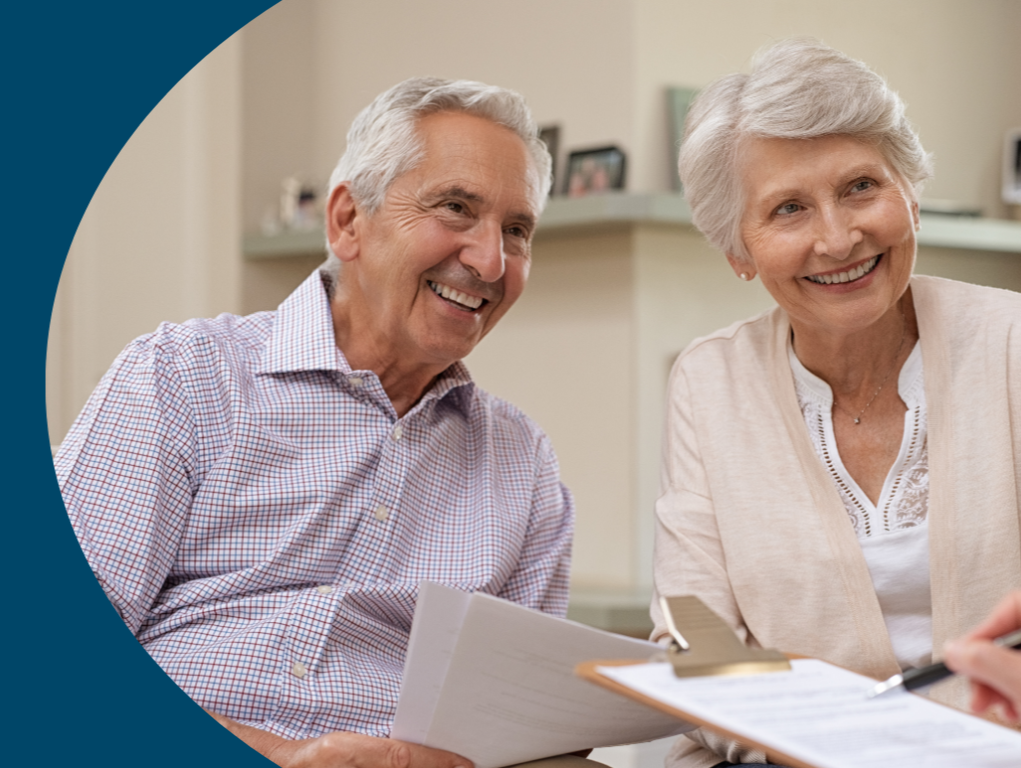 An elderly couple sitting at a table, smiling and looking at a document, with a clip on it, in a cozy home setting.