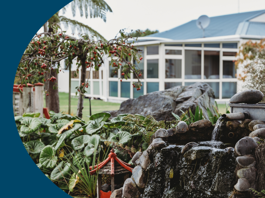 A garden with a water fountain, surrounded by plants and trees, with a white house or greenhouse in the background.