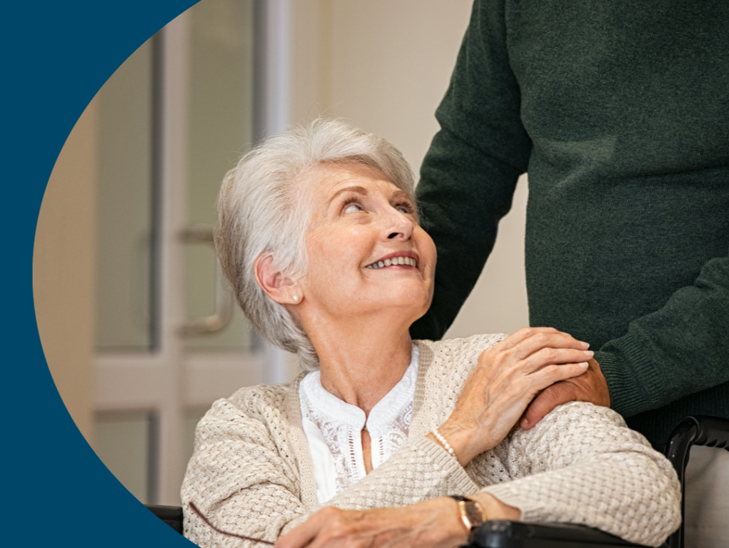 An elderly woman with white hair smiling and looking up, sitting in a wheelchair, while a person standing next to her places a hand on her shoulder.