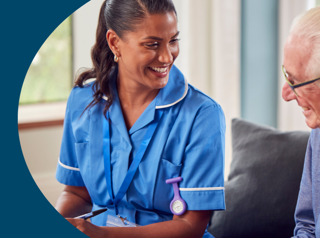A female nurse in blue scrubs smiling and talking to an elderly man in a gray sweater.