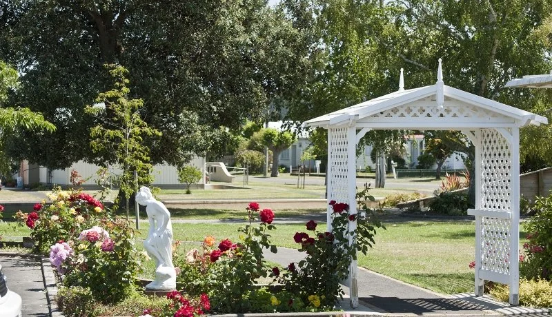 White garden gazebo surrounded by colorful roses and greenery in a park-like setting.