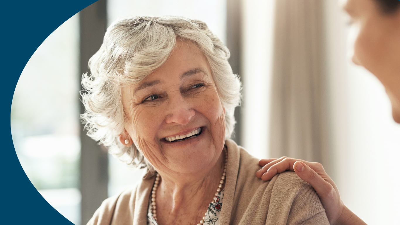 An elderly woman smiling warmly as she talks to a caregiver who has their hand on her shoulder.