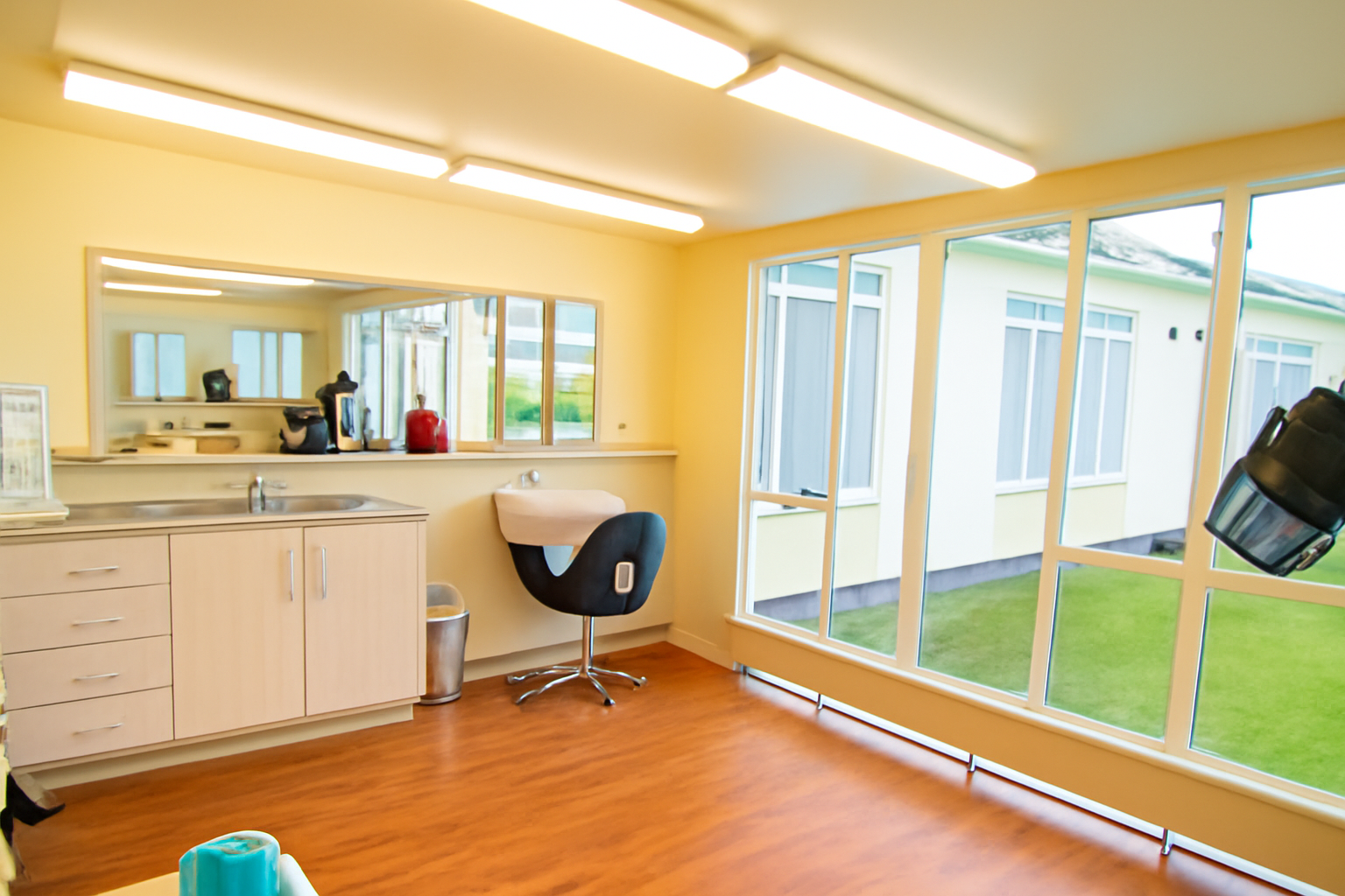 Indoor room with large windows showing a grassy outdoor area, a black chair in front of a small sink and cabinet, a mirror behind, and a black hair dryer mounted on the wall to the right.