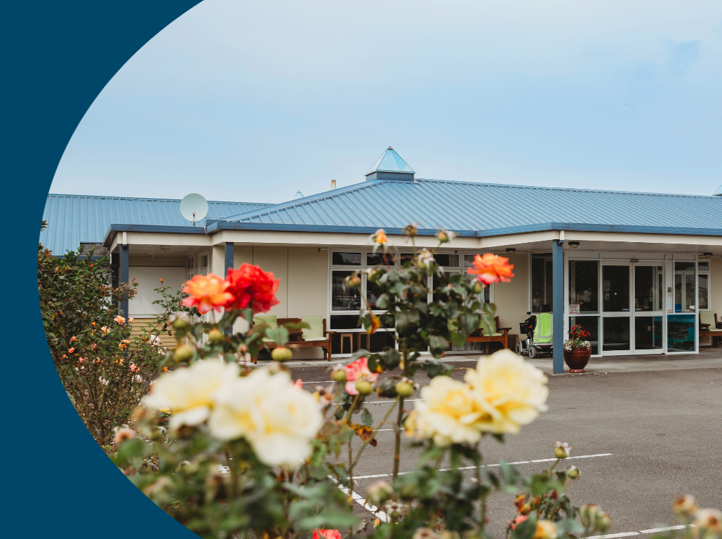 A building with a blue metal roof surrounded by a parking lot and colorful flowers in the foreground.
